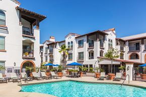 Laguna Woods apartment for rent - A large swimming pool is surrounded by lounge chairs and umbrellas in front of a row of apartment buildings.