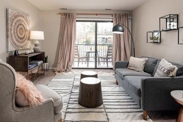 Henrico apartment for rent - A living room with a grey couch, a grey armchair, a wooden side table with a lamp, a round wooden table, and a large window with curtains.