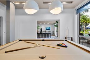 Hallandale Beach apartment for rent - A pool table with a cue stick and balls is in the foreground of a room with a view of a dining area and outdoor patio.
