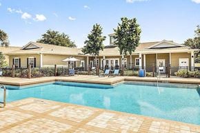 Bradenton apartment for rent - Community pool with sundeck, lounge chairs, surrounded by black metal fence with trees, lake, and exterior of clubhouse in the background