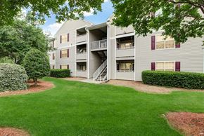 Durham apartment for rent - An apartment building with gray siding and burgundy shutters, is three stories tall with a balcony/patio on each level, and has stairway access in the middle of the building. The building is surrounded by grass and trees.