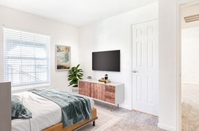 Federal Heights apartment for rent - Model Bedroom with Bed Next to Window Next to Framed Wall Art Next to Plant, Dresser and Mounted TV