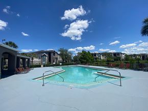 Orlando apartment for rent - Community pool with sundeck, lounge chairs, surrounded by black metal fence with trees, lake, and building exteriors in the background