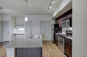 Burleson apartment for rent - Kitchen with island, dark wood cabinetry, stainless appliances with wood style flooring, granite countertop, track lighting, and built-in desk in background