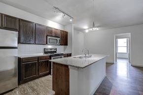Burleson apartment for rent - Open kitchen with wood style flooring, dark wood cabinetry, stainless appliances, granite countertops, double basin sink, track lighting and hanging bar top light