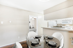 Plano apartment for rent - Virtually staged dining room with round table and chairs, wood style flooring, hanging light fixture, large window with blinds, and kitchen with breakfast bar in background