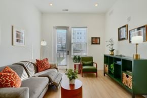 Baltimore apartment for rent - Now leasing senior 1-bedrooms in Baltimore, MD. A living room with a grey couch, a green chair, and a green bookshelf.