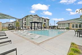 Manor apartment for rent - A large swimming pool surrounded by lounge chairs and umbrellas in front of a multi-story apartment building.