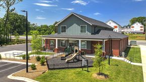 Charlotte apartment for rent - Exterior view of a community clubhouse and playground area, featuring a brick and siding building with a covered porch, fenced play structure with slides, and landscaped grounds.