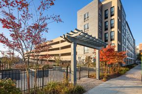 Wyandanch apartment for rent - A parking lot is surrounded by a black fence and a tree with red leaves.