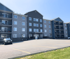 Fredericton apartment for rent - A black car is parked in a parking lot in front of a large apartment building.