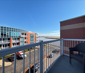 Moncton apartment for rent - A balcony with a chair and a railing overlooking a parking lot and buildings.