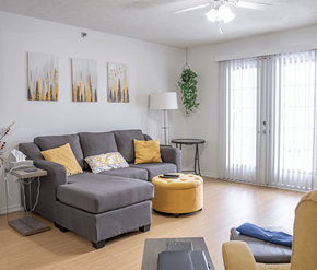 Moncton apartment for rent - A living room with a grey couch, yellow ottoman, and a ceiling fan.