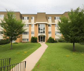 Fredericton apartment for rent - A building with a brown roof and tan walls with a walkway in front.