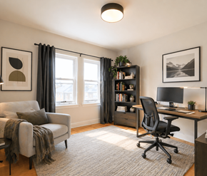 Moncton apartment for rent - A cozy room with a grey couch, a black chair, a desk with a computer, and a bookshelf with books and plants.