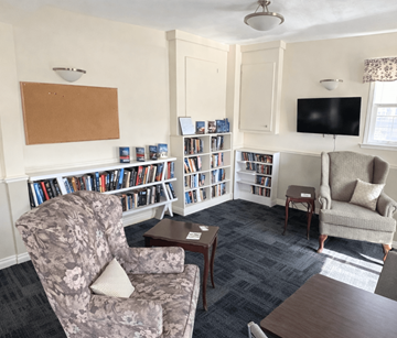 Fredericton apartment for rent - A living room with a grey floral armchair, a wooden coffee table, a grey sofa, a bookshelf with books, a television, and a window with curtains.