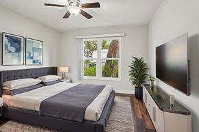 Inglewood apartment for rent - A bedroom with a bed, a ceiling fan, a window with a view of trees, a nightstand with a lamp, a plant, and a flat-screen TV mounted on the wall.