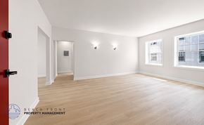 Los Angeles apartment for rent - A spacious room with wooden flooring and white walls, featuring a red door and a window with a view of a building outside.
