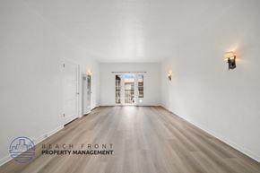 Riverside apartment for rent - A spacious room with wooden floors and white walls, with a logo for Beach Front Property Management in the bottom left corner.
