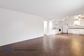 Long Beach apartment for rent - A spacious, empty room with wooden floors and white walls, with a logo for Beach Front Property Management in the bottom left corner.