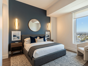 Oakland apartment for rent - A bedroom with a large bed, a round mirror, and a window overlooking a cityscape.