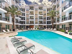Houston apartment for rent - A large swimming pool surrounded by lounge chairs and palm trees in front of a multi-story apartment building.