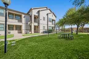 Waco apartment for rent - A sunny day at a grassy park with a bench and apartment buildings in the background.