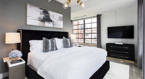 Atlanta apartment for rent - A bedroom with a large bed, a nightstand with a lamp, a side table with a vase of flowers, a TV, and a window with a view of the city.