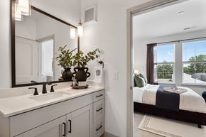 Carmel apartment for rent - A view from a modern bathroom vanity featuring a light wood cabinet and quartz countertop, looking through an open doorway into a bright, carpeted bedroom with large windows.