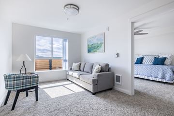 Portland apartment for rent - A well-lit, modern living room with a grey sofa, a small table with a lamp, and a bed with a blue and white comforter in the background.