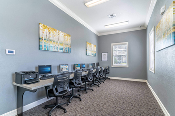 A room with grey walls and a carpeted floor has a row of chairs and desks with computers on them.