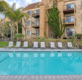 A pool in front of a tan building with trees and chairs around it.