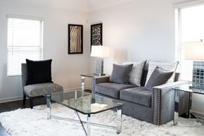 Humble townhouse for rent - Two-Bedroom Apartments in Humble, TX - Edgewater at the Lagoon - Living Room with Vinyl Plank Flooring, Fluffy White Area Rug, Glass Cofee Table, End Tables with Lamps, Grey Couch, Graphic Cushioned Chair, Two Windows, and Framed Wall Art.