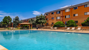 Aurora apartment for rent - A large swimming pool in front of a brown building with a fence and trees.