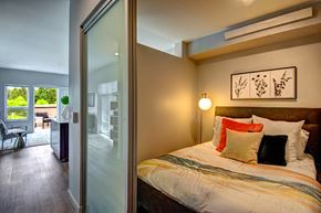Bothell apartment for rent - Bothell Apartments - 98th Avenue - A Bedroom With A Sliding Frosted Glass Door, Layered Pillows, Floor Lamp, And Wood-Style Flooring.