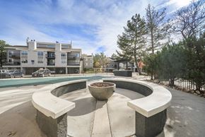 Aurora apartment for rent - Apartments in Aurora, CO, with an In-Unit Washer and Dryer - The Juliet at Lowry - A Fire Pit Surrounded by a Circular Concrete Bench, Metal Fence, Trees, and a View of the Parked Cars and Apartments.