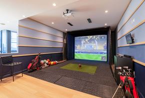 New Rochelle apartment for rent - The indoor golf simulator room at Encore 325 Apartments features a projector screen displaying a green course, a striking mat, a golf bag, and blue padded walls.
