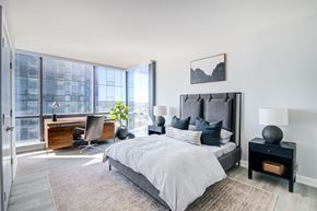 New Rochelle apartment for rent - A luxurious and bright bedroom features a gray upholstered bed, a large corner window with a city view, and an abstract black and white painting above the headboard.