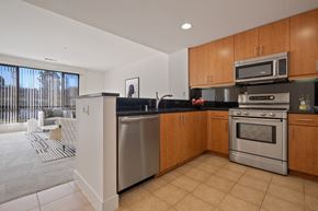 Novato apartment for rent - Dog-Friendly Apartments in Novato, CA - Millworks - Kitchen With Wooden Cabinets, Stainless Steel Appliances, Black Countertops, Tile Flooring, And An Adjacent Living Room With Large Windows.
