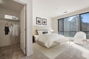 Novato apartment for rent - Novato, CA, Pet-Friendly Apartments - Millworks - Bedroom With White Bedding, Spacious Closet, Large Window With Vertical Blinds, White Accent Chair, Bench, And Carpeted Flooring.