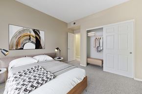 Upland apartment for rent - Dog-Friendly Apartments in Upland, CA - Park Central Apartments - Bedroom with Carpet Flooring, Large Bed, Nightstands, Lamps, Abstract Wall Art, and Oversized Closets with a Sliding Door.
