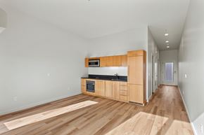 Portland apartment for rent - North East Portland, OR, Pet-Friendly Apartments - The Beverly - Open Apartment Kitchen And Living Area With Wood Style Flooring, Light Wood Cabinets, Black Countertops, And Bright Hallway.