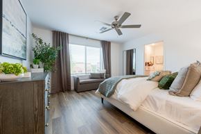 Tolleson apartment for rent - Townhomes in Tolleson, AZ, With In-Unit Washer and Dryer - Villas at 91st - Bedroom With Plush Bed, Ceiling Fan, Large Window With Curtains, Wood-Style Flooring, Dresser, Painting, and Bed Bench.