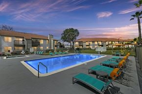 Moreno Valley apartment for rent - Apartments for Rent in Moreno Valley, CA - Ardella on Chagall - Dusk View of Gated Swimming Pool with Spa, Lounge Seats, Tables and Chairs with Umbrellas, and Apartment Buildings in the Background.