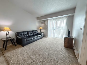 Williston apartment for rent - A black leather couch sits in a living room with a carpeted floor and a window with blinds.