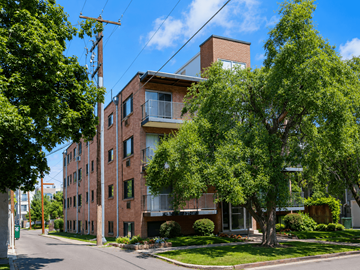 Denver apartment for rent - A red brick building with a balcony on the second floor is surrounded by green trees.