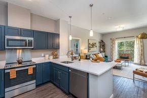 Lowell apartment for rent - The Residences at Crosspoint open-plan kitchen and living area includes navy blue shaker cabinets, stainless steel appliances, a white island countertop with stools, and seating near a sliding glass door leading to a balcony.