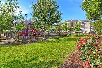 A playground with a red slide and a purple canopy is surrounded by a green lawn and a fence