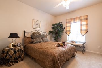 Overland Park apartment for rent - A bedroom with a bed, a nightstand with a lamp, a window with curtains, and a plant.