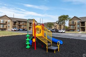 Olathe apartment for rent - Playground with slide, swings, and seesaw, with apartment buildings in the background.